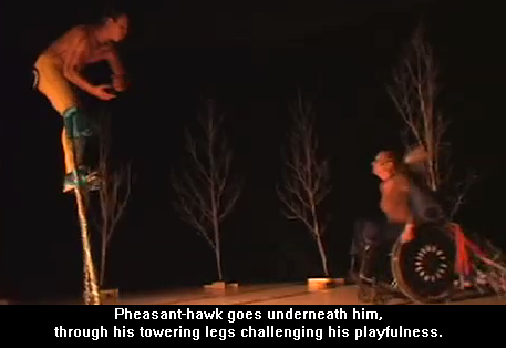 A screen capture from a video of a performance of 'The Meeting' in which a tall walking animal played by an actor on stilts meets an short rolling animal played by an actress using a wheelchair. The caption reads 'Pheasant-hawk goes underneath him, through his towering legs challenging his playfulness'.